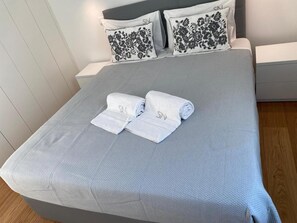 Un letto rifatto con cura, coperto da una coperta grigia, due cuscini con motivi floreali neri e due asciugamani arrotolati con un monogramma.