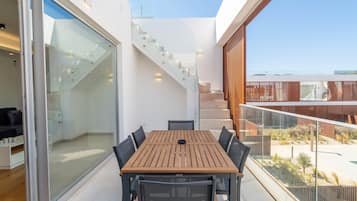 Ein moderner Essbereich im Freien mit einem Holztisch und Stühlen, einer Glastür, die zu einer Treppe führt, und Blick auf die umliegende Landschaft.