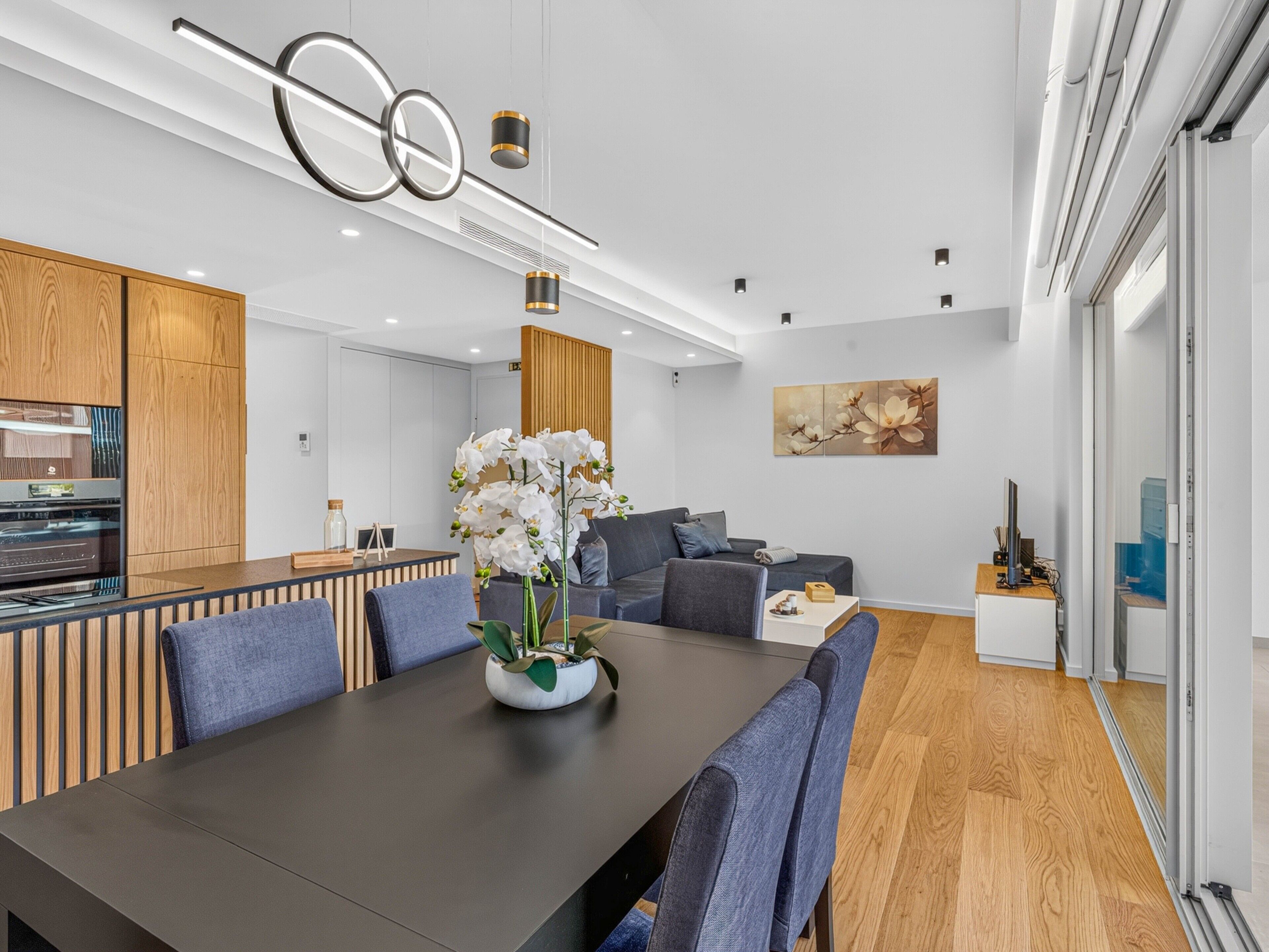 Una cocina moderna con armarios de madera, un comedor con mesa oscura y sillas azules, y una sala de estar con sofá y una mesa de centro.