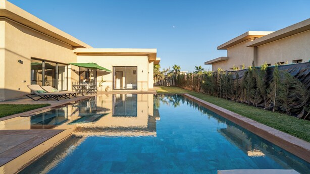 Piscina con sillones de descanso y zona de estar cubierta, rodeada por una valla baja y vegetación.