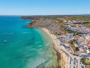 An aerial view of a coastal town with a beach, clear blue waters, and buildings along the shore.