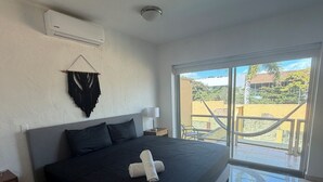 Une chambre avec un grand lit, un climatiseur fixé au mur, une fenêtre donnant sur l’extérieur et une petite table de chevet.