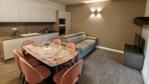 A modern kitchen with white cabinets, a built-in oven, and a microwave. There is a dining table with pink chairs and a marble top. A grey sofa is against the wall, and a TV is mounted on the wall. - Modern and cozy suite for couples and families "Calliope" (Vernazzano Basso)