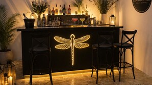 A bar area with a dragonfly light, two bar stools, a white pergola, and a wall-mounted sign for Dragonfly Dream Weddings. - Dragonfly Retreat Málaga | Private Villa & Rose Pool (Coín)