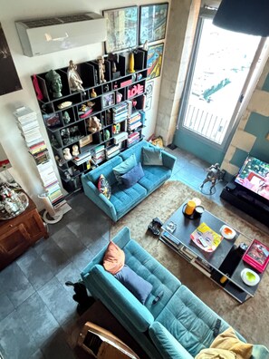 A living room with two blue sofas, a coffee table, a bookshelf filled with books and figurines, and a large window with a view. - Exceptional duplex loft on Marseille's Old Port - Panoramic view (Marseille)