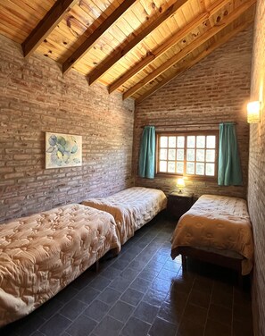 A room with two beds, a brick wall, a wooden ceiling, a window with curtains, and a painting on the wall.