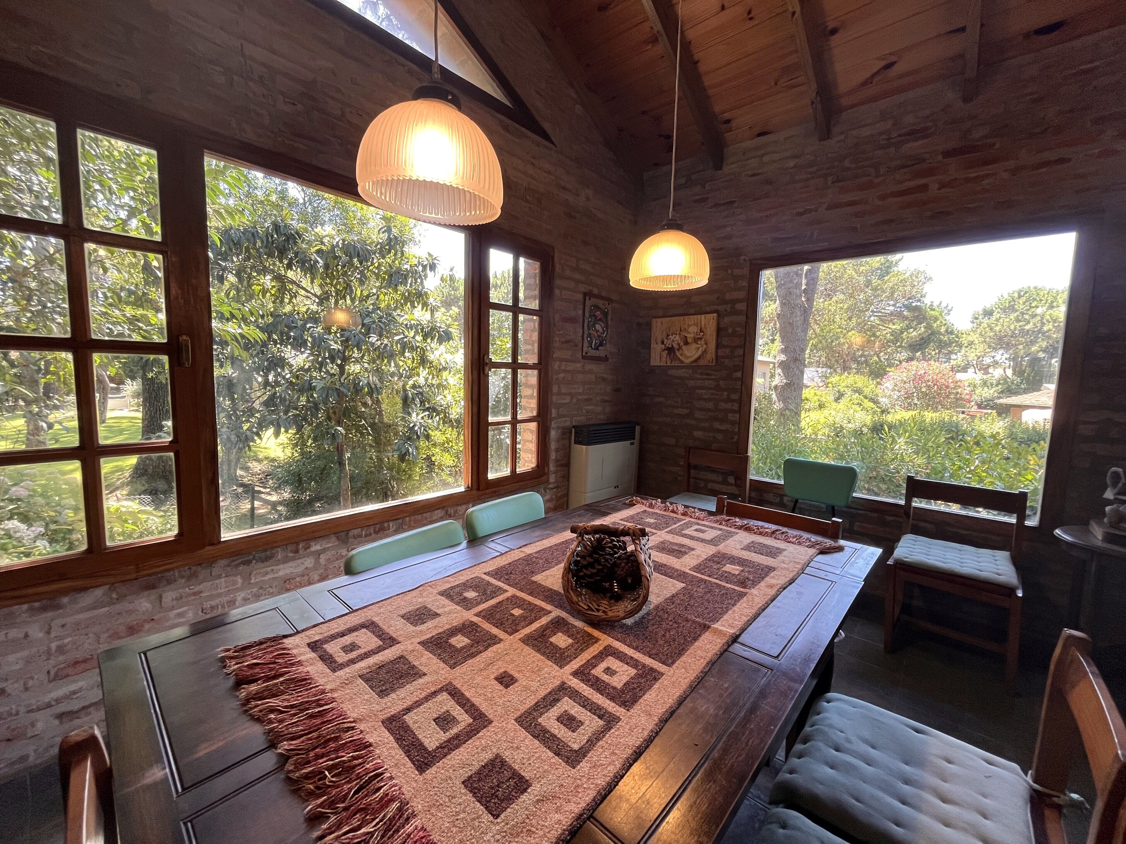 A rustic dining area with a patterned tablecloth, wooden chairs, and large windows with a view of greenery.