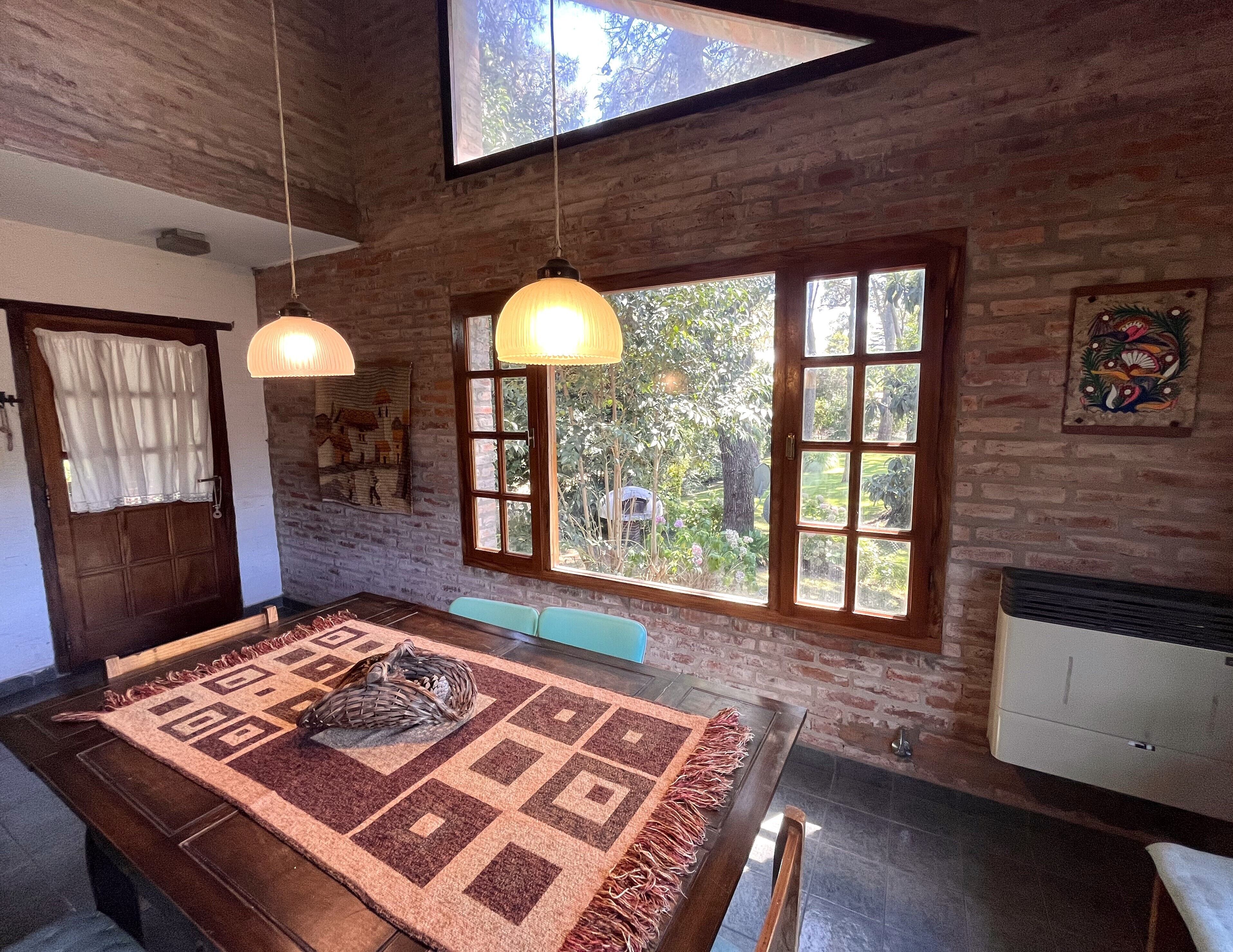 A dining area with a wooden table, patterned rug, hanging pendant lights, and large windows with a view of greenery.