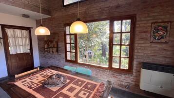 A dining area with a wooden table, patterned rug, hanging pendant lights, and large windows with a view of greenery.