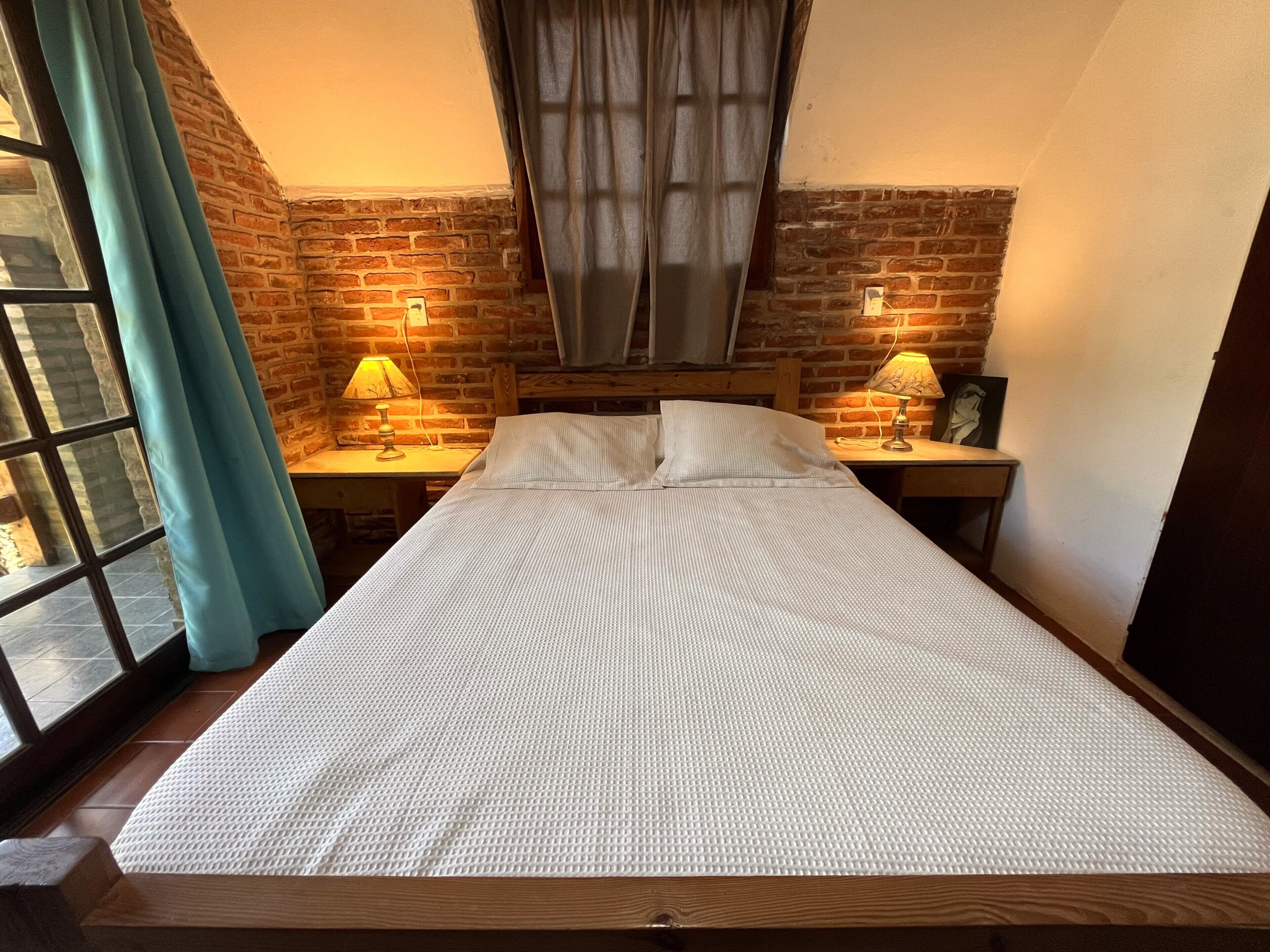 Un lit simple avec un matelas blanc texturé, deux lampes de chevet, une tête de lit en bois, un mur en briques et une fenêtre avec des rideaux.