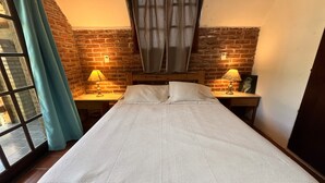 Un lit simple avec un matelas blanc texturé, deux lampes de chevet, une tête de lit en bois, un mur en briques et une fenêtre avec des rideaux.