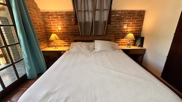 Un lit simple avec un matelas blanc texturé, deux lampes de chevet, une tête de lit en bois, un mur en briques et une fenêtre avec des rideaux.