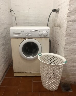 Une machine à laver de la marque Candy, de couleur blanche, avec un tambour à chargement frontal, un panier à linge blanc avec une anse verte et un sol carrelé.