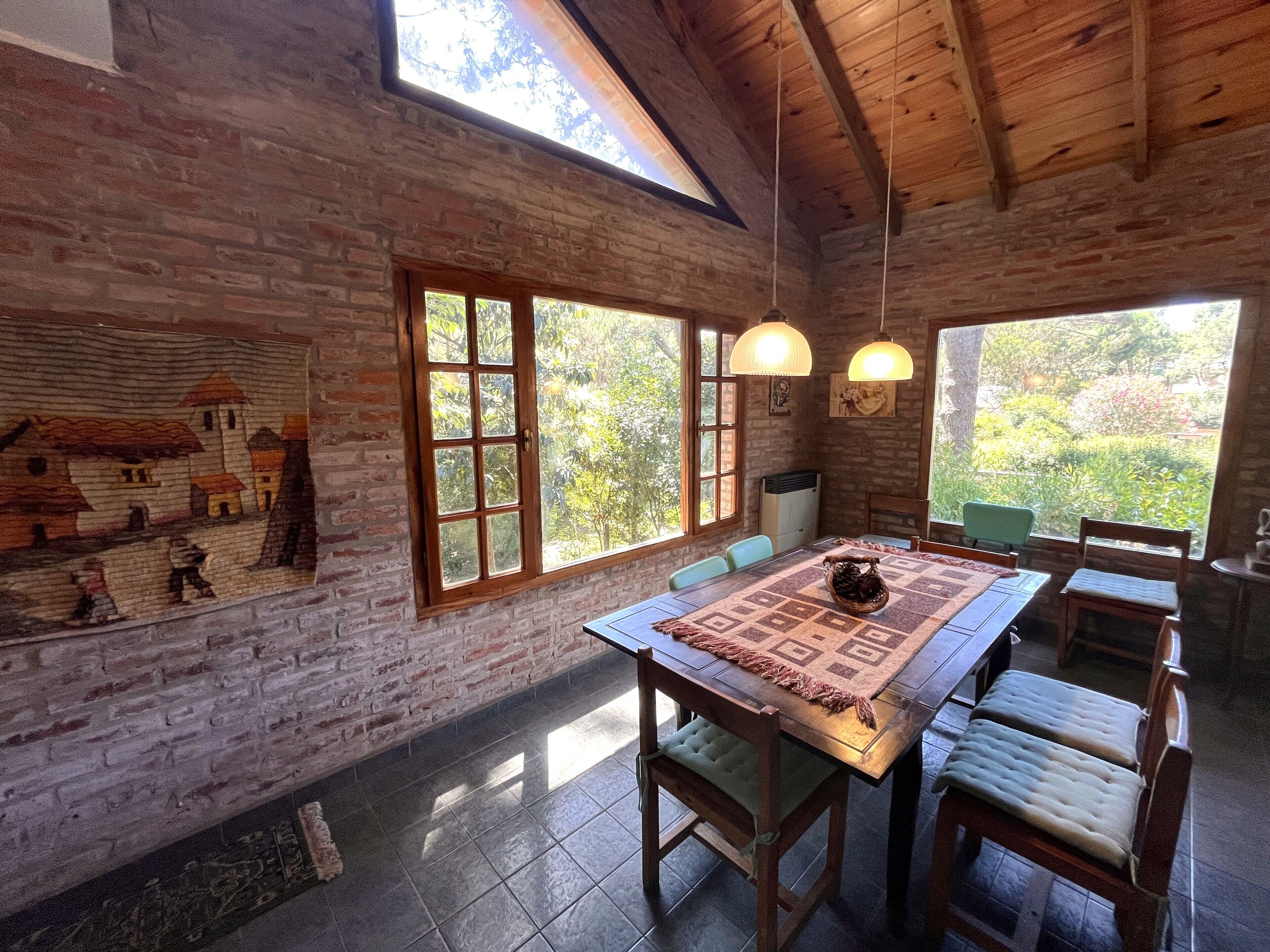 A rustic dining area with a long table, chairs, and a view of greenery through large windows.