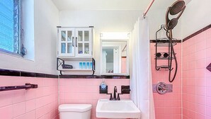 Ein Badezimmer mit rosa gefliesten Wänden, Toilette, Waschbecken, einem Schrank mit Pflegeprodukten und einer Dusche inklusive Handbrause.