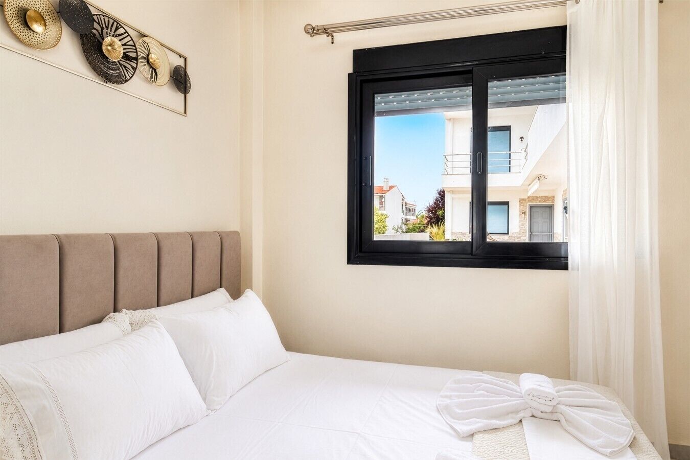 Cama bem arrumada com lençóis brancos, cabeceira e janela com vista para edifícios.