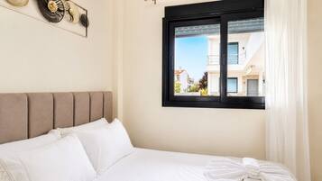 Cama bem arrumada com lençóis brancos, cabeceira e janela com vista para edifícios.