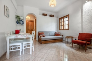 Un salon comprenant une table à manger, un canapé, un fauteuil rouge, une porte en bois et une fenêtre.