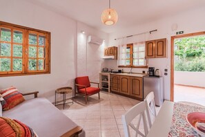 Une pièce de taille modeste comprenant un lit, une chaise rouge, une petite table, un coin cuisine et une vue sur la verdure.