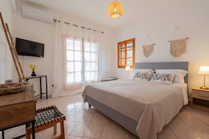 Une chambre à coucher comprenant un lit, une télévision, une fenêtre, une lampe et une décoration murale en macramé.