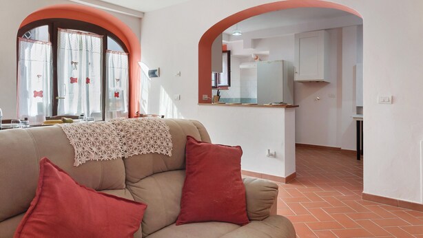 A living room with a sofa, red cushions, and a lace throw. There is a kitchen visible through an archway with white cabinets and a countertop. - Casa del Custode: Via degli Dei – Mugello Circuit (Tagliaferro)