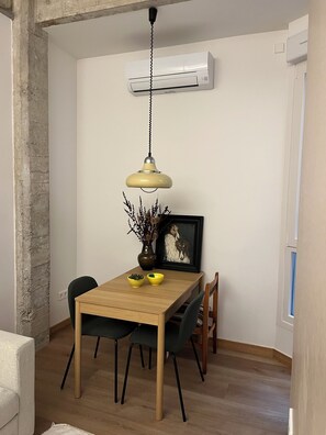 Een eetgedeelte met een houten tafel, twee stoelen, een hanglamp, een vaas met gedroogde planten en een lijst met een afbeelding.