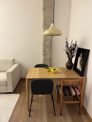 Un área de comedor con una mesa de madera, dos sillas, una lámpara colgante, un jarrón con plantas secas y un cuadro en la pared.