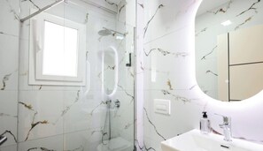 Ein Badezimmer mit einer gläsernen Duschkabine, einer weißen Marmorwand, einem runden Spiegel und einem weißen Waschbecken mit Seifenspender.