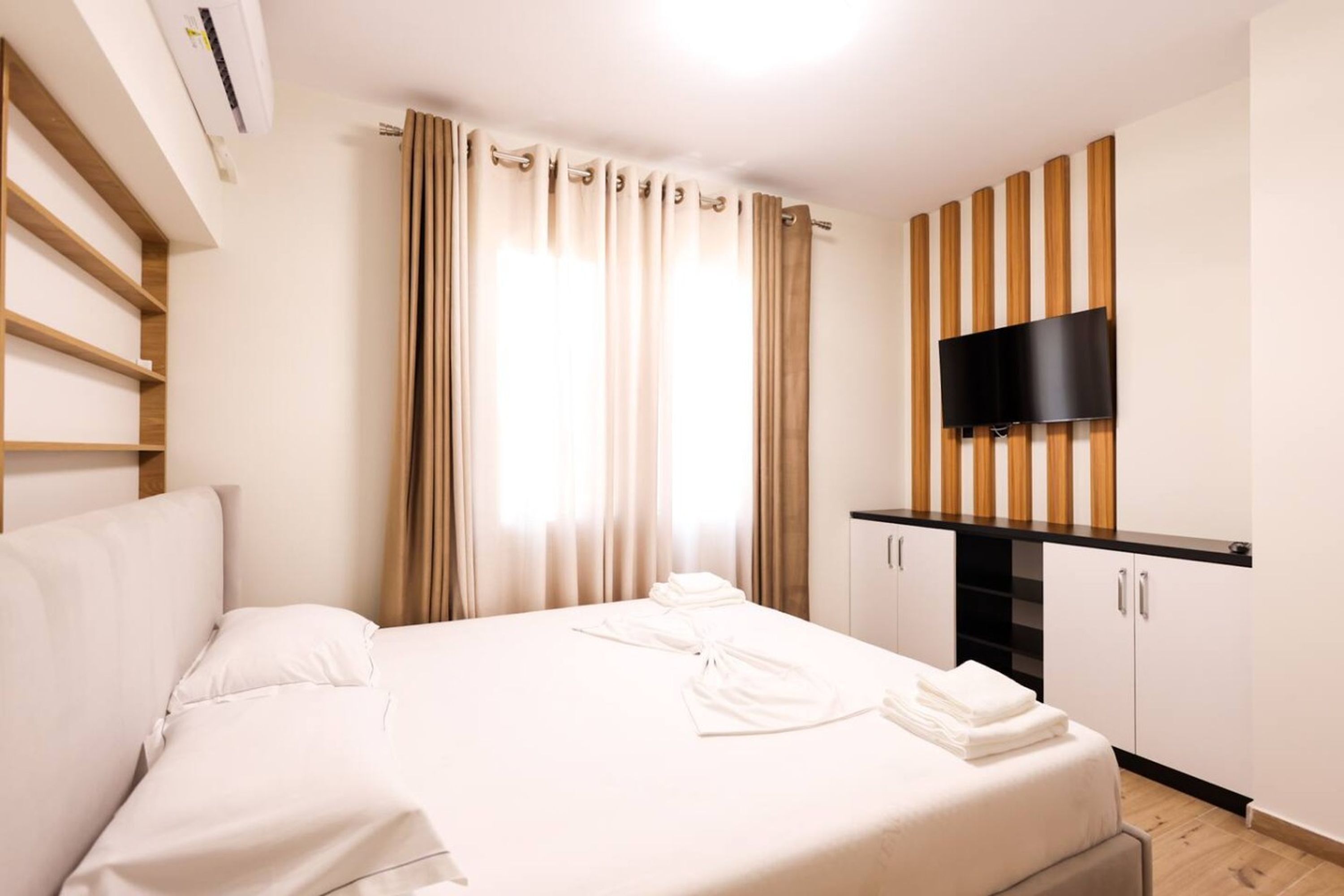 Ein Hotelzimmer mit Bett, Flachbildfernseher an einer Holzverkleidung, Kleiderschrank und einem Fenster mit Vorhängen.