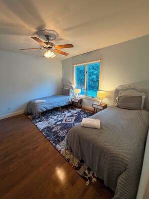 Une chambre avec deux lits, un ventilateur de plafond, une fenêtre avec vue, un tapis à motifs et une petite table avec une lampe.