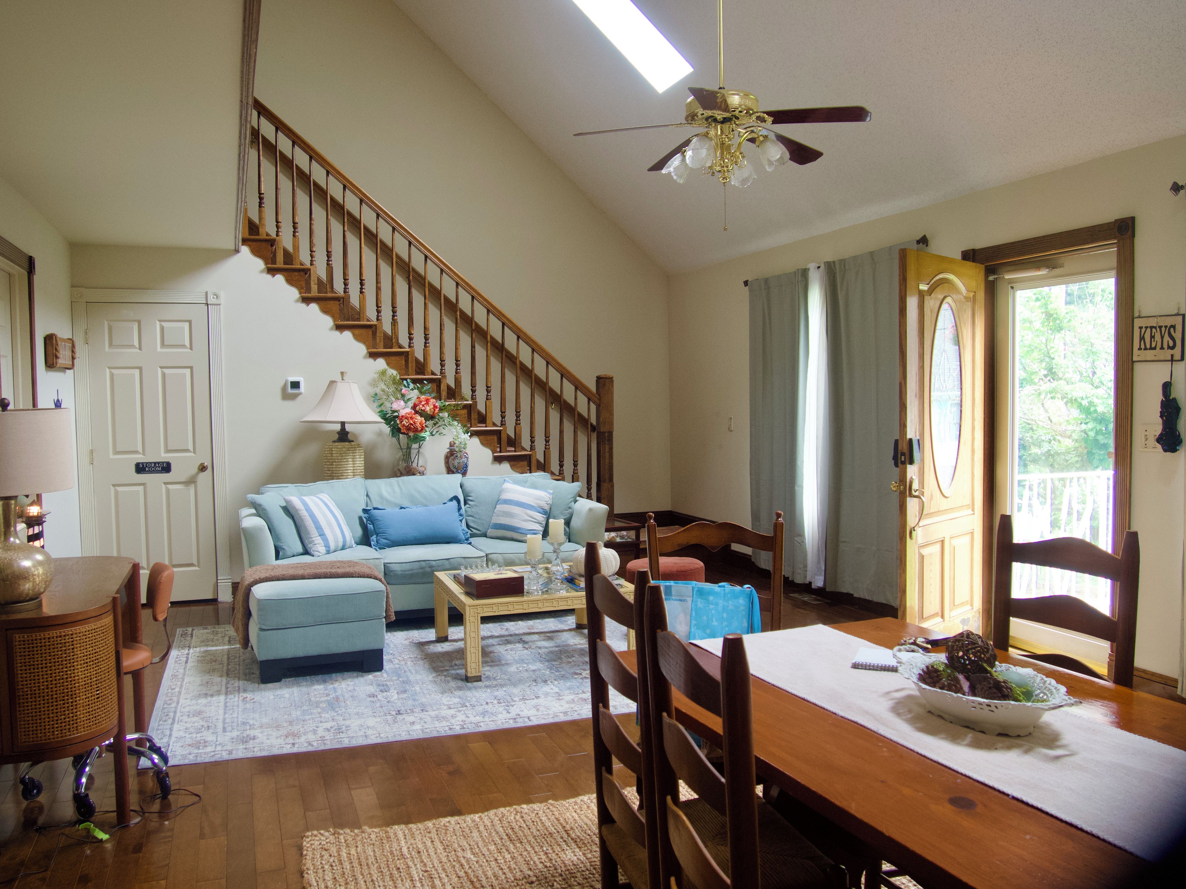 A living room with a staircase, a dining table with chairs, a sofa, and a ceiling fan.