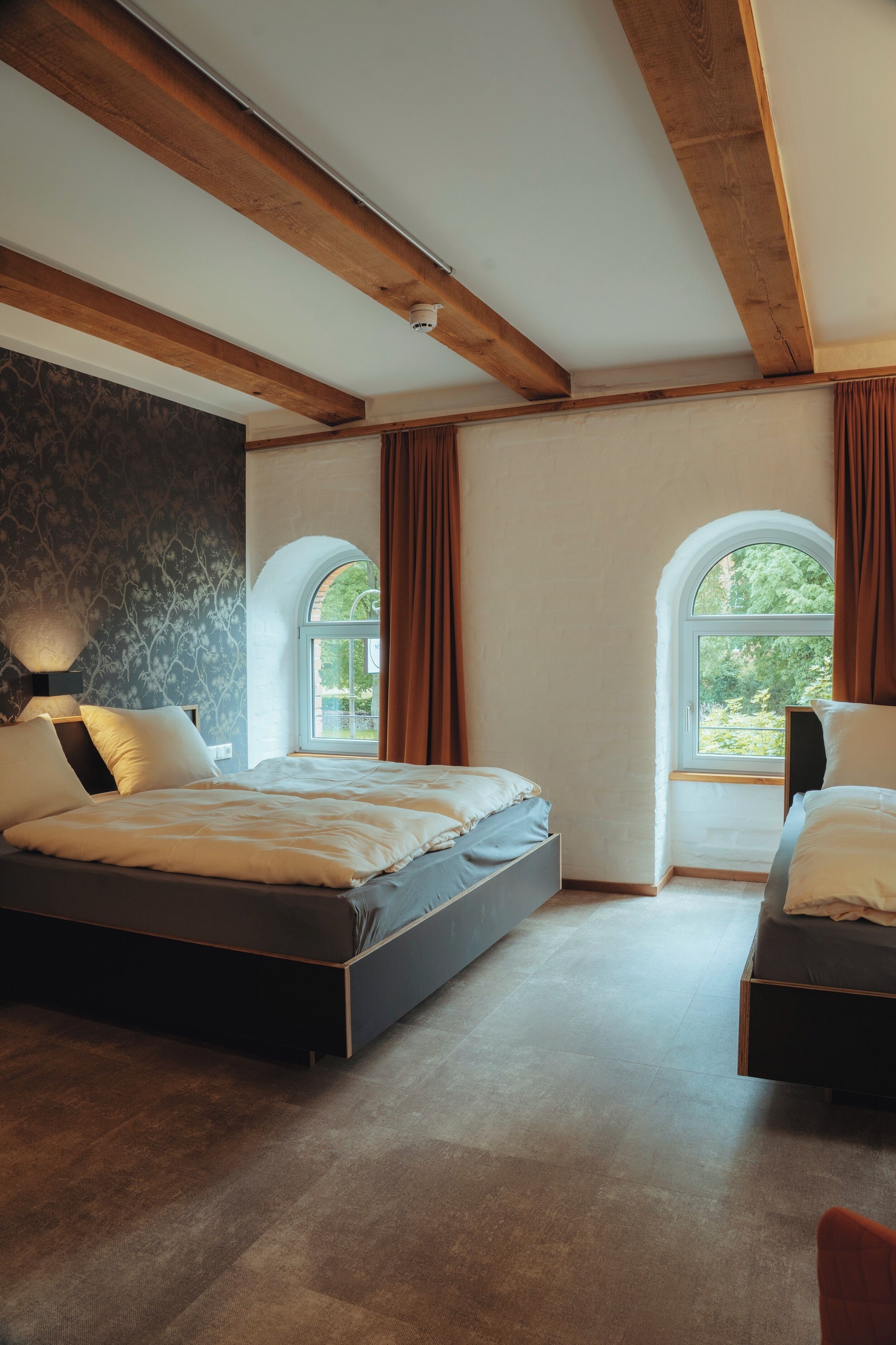 Un dormitorio con una cama grande, dos camas más pequeñas, una ventana con cortinas y un techo con vigas a la vista.