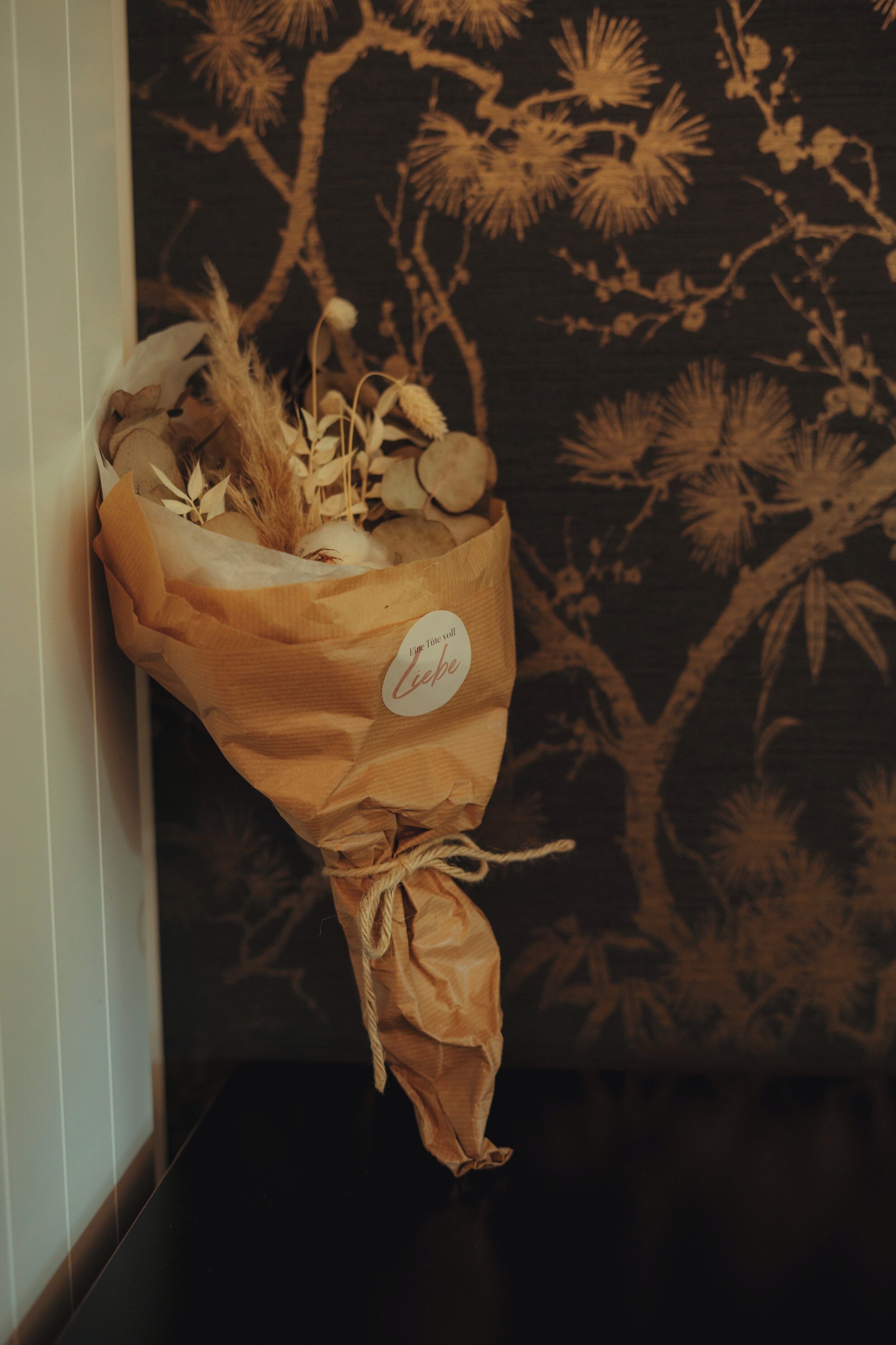 Un bouquet de fleurs, attaché avec une ficelle, est posé devant un mur recouvert d’un papier peint à motifs.