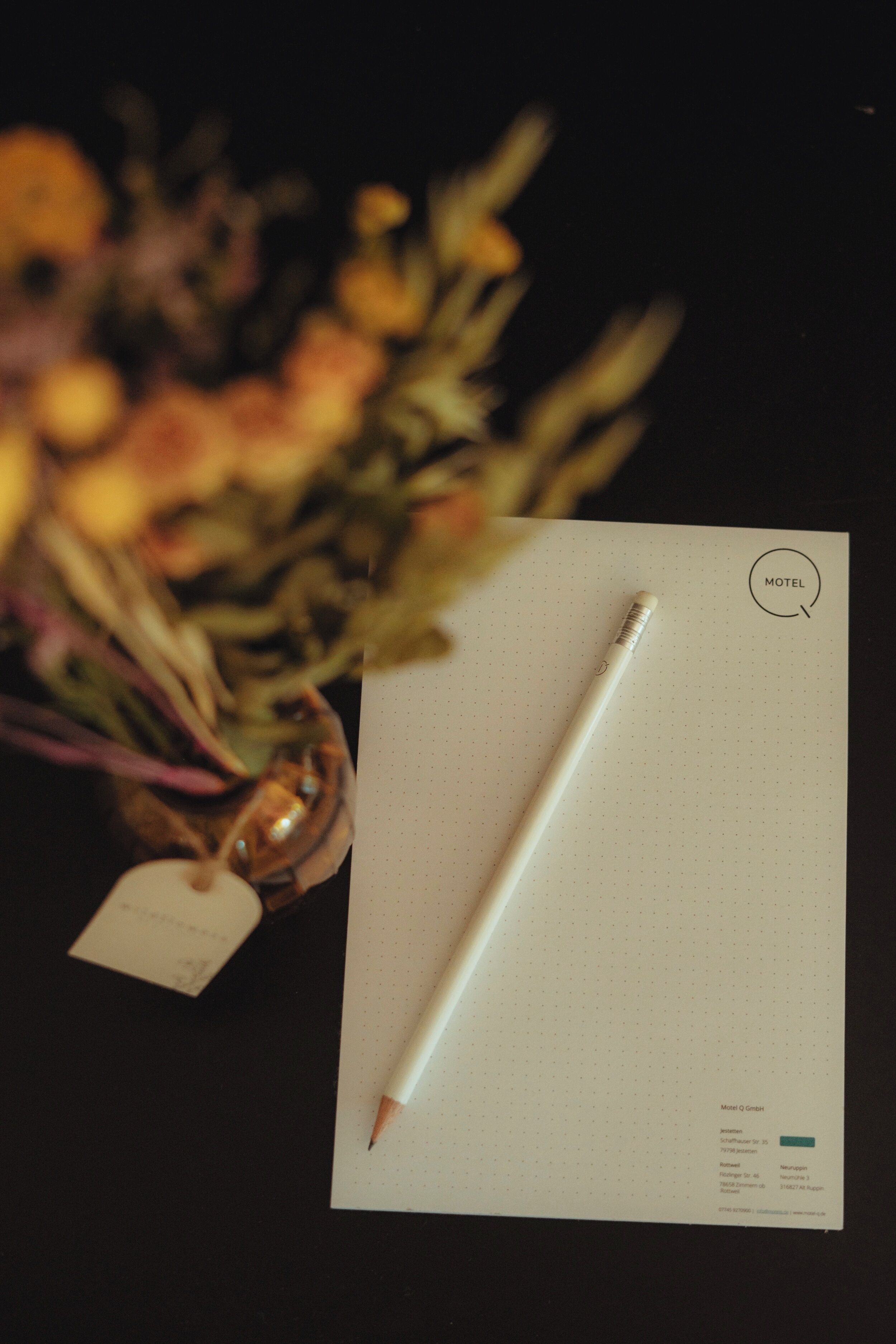 A white notepad with a pencil, a tag, and a small bouquet of flowers on a dark surface.