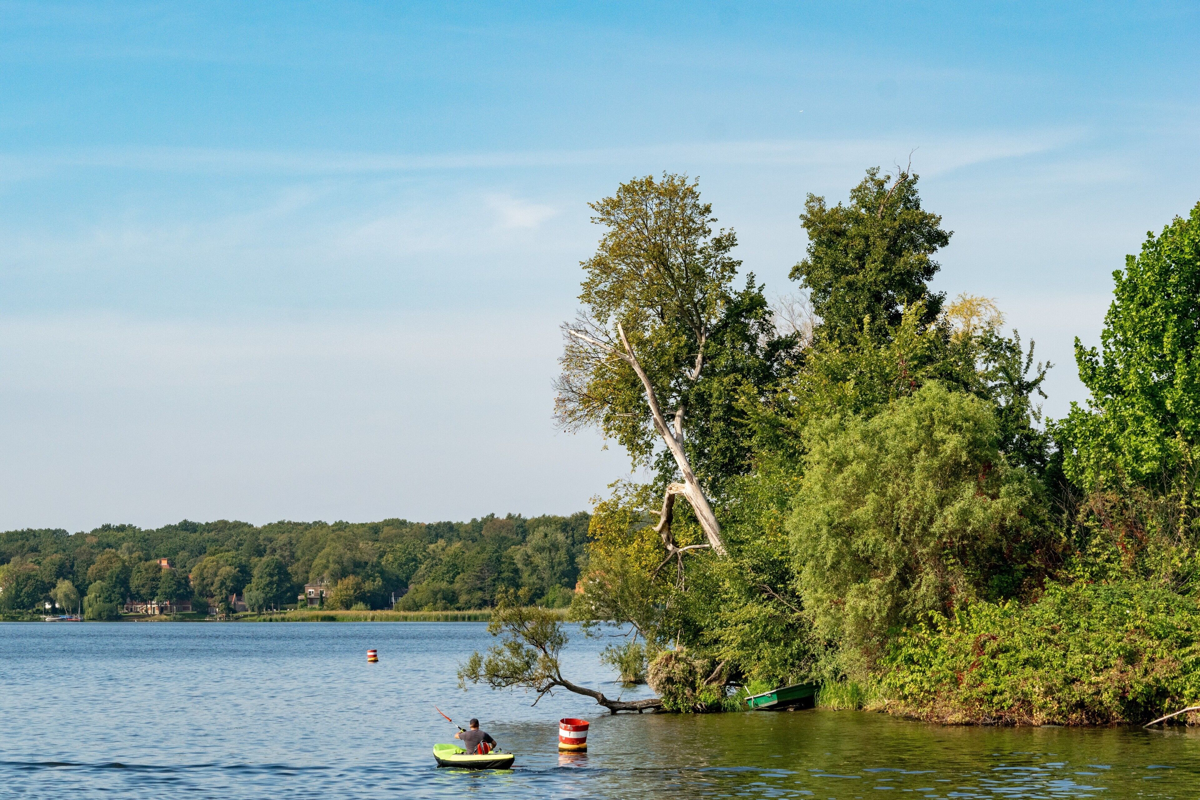 A calm lake with a single kayak and a buoy, surrounded by dense trees and a clear sky.