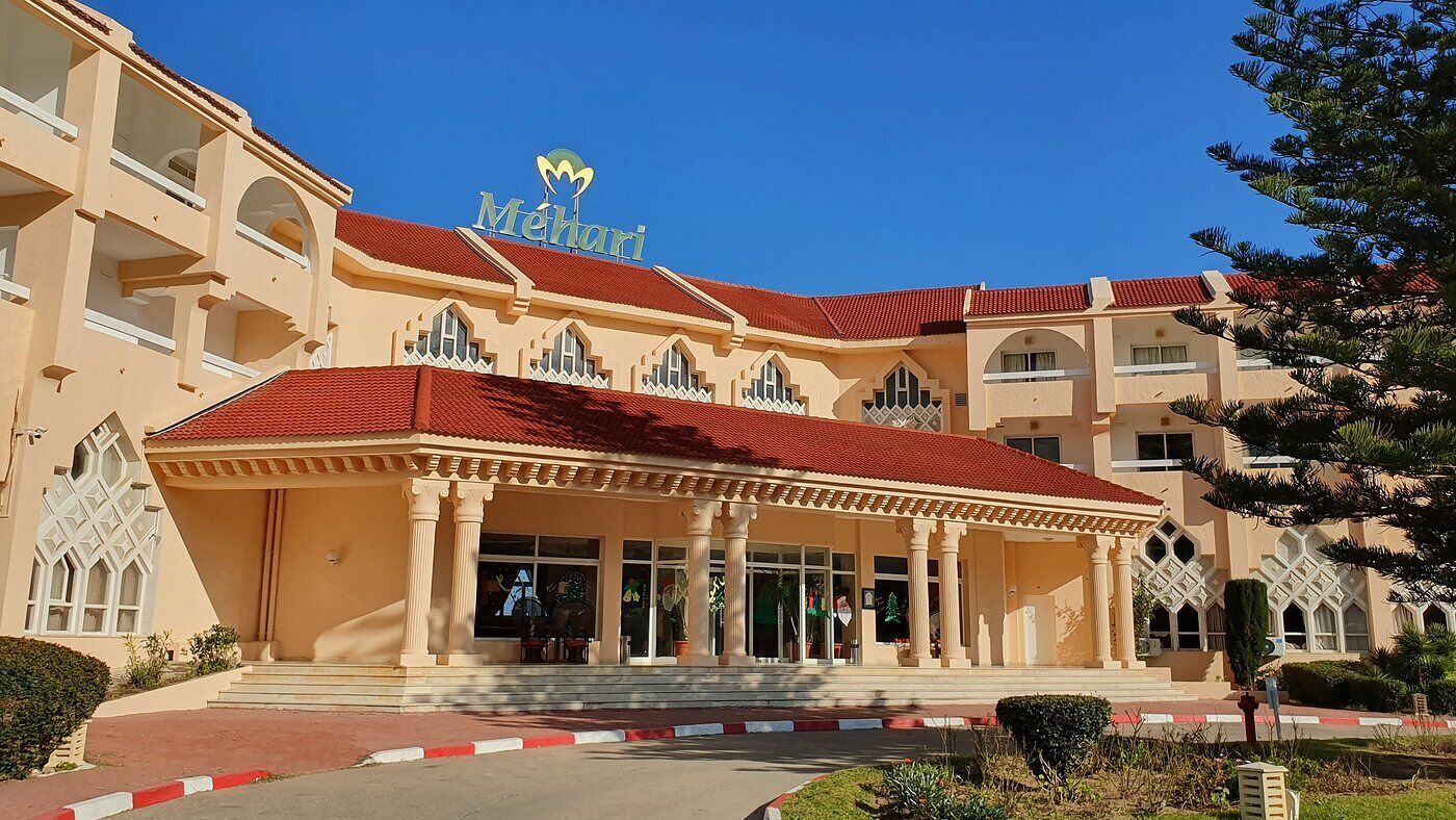 Un hotel con techo de tejas rojas, un acceso cubierto con columnas y un letrero con el logotipo estilizado de la letra 'M'.