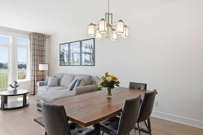 A modern living room with a sofa, dining table, chairs, a chandelier, and a vase of flowers. - Ski & Tee Retreat - Walk to Village (The Blue Mountains)