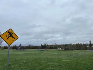 A pedestrian crossing sign in a grassy field with playground equipment and a gazebo in the background.