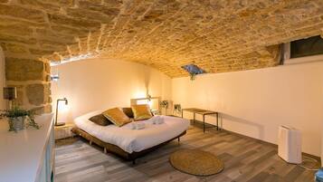A bedroom with a stone ceiling, a bed with pillows, a bedside table, a small table, and a radiator.