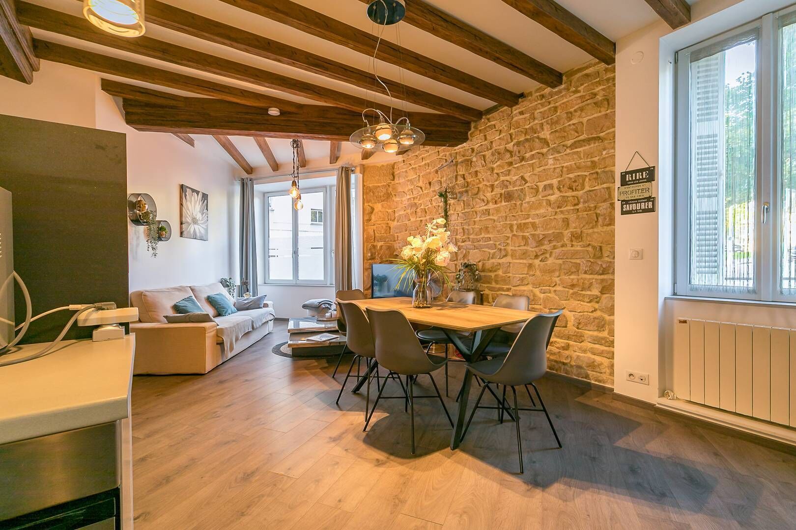 Un salon chaleureux avec un mur en pierre, des poutres apparentes en bois au plafond, une table à manger avec des chaises et un canapé.