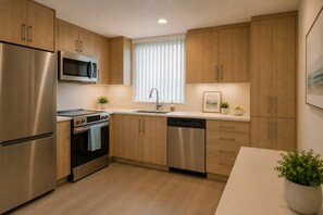 Eine moderne Küche mit Edelstahlgeräten, Holzschränken, einem Geschirrspüler und einem Fenster mit Jalousien.