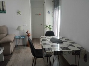 Ein kompakter Wohnraum mit Esstisch, Stühlen, einer Couch, einem kleinen Beistelltisch mit Lampe und einer Tür mit dem Aufdruck 'PRIVADO'.