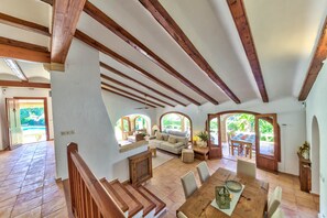 A spacious living area with a high ceiling, wooden beams, a sofa set, a dining table, and a view through arched windows. - Casa Violeta — Mediterranean Art Villa in Jávea (Xàbia)
