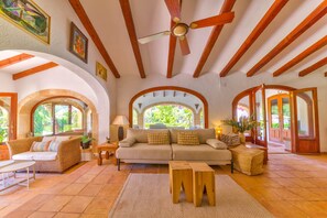 A spacious living area with a high ceiling, wooden beams, a ceiling fan, and large windows offering views of greenery. - Casa Violeta — Mediterranean Art Villa in Jávea (Xàbia)
