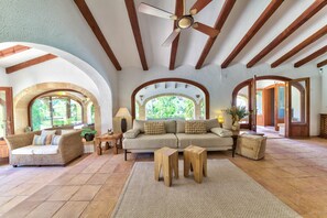 A spacious living room with a sofa, two wooden stools, a ceiling fan, and arched windows. - Casa Violeta — Mediterranean Art Villa in Jávea (Xàbia)