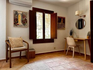 Ein Zimmer mit Schreibtisch aus Holz, zwei Stühlen, einem Fenster mit geöffneten Klappläden und einem an der Wand befestigten Kunstwerk.