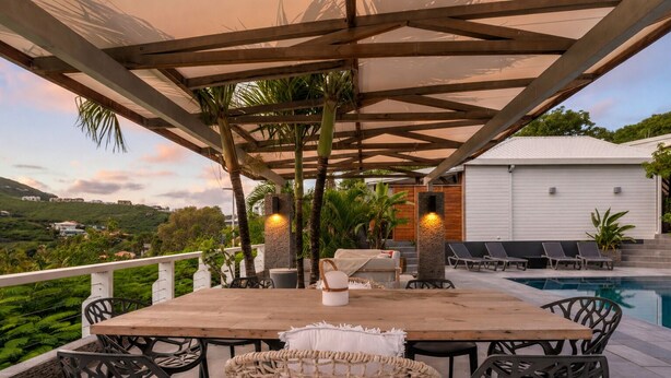 An outdoor patio with a pool, a wooden table, wicker chairs, and a white building in the background. - NAYA (Saint-Martin)