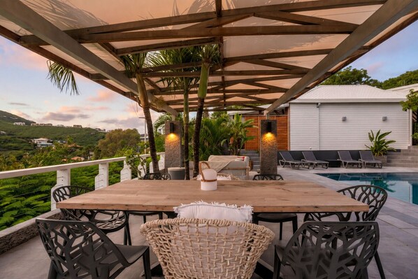 An outdoor patio with a pool, a wooden table, wicker chairs, and a white building in the background. - NAYA (Saint-Martin)