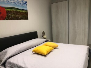 Ein ordentlich bezogenes Bett mit einem gemusterten Bettbezug und zwei Kissen, ein an der Wand befestigtes Kunstwerk sowie ein Kleiderschrank.