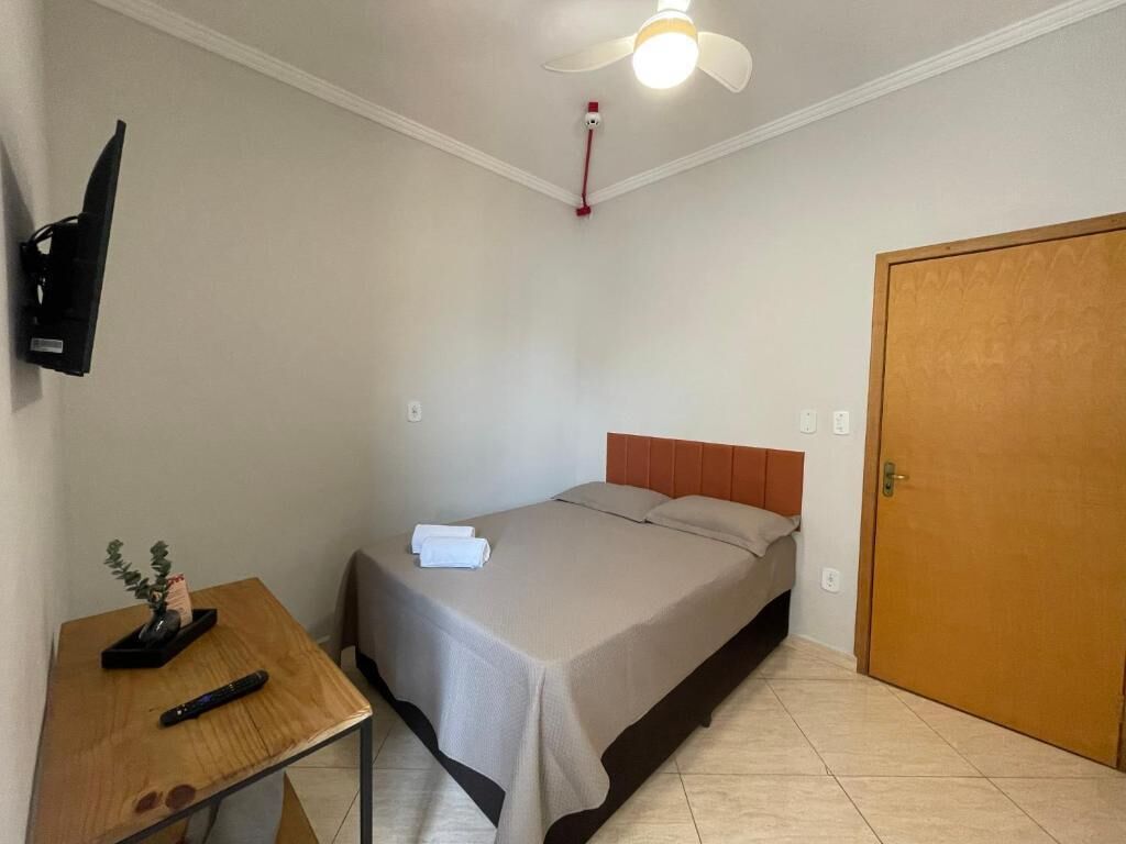 A bedroom with a bed, a desk with a remote control, a ceiling fan, and a door.
